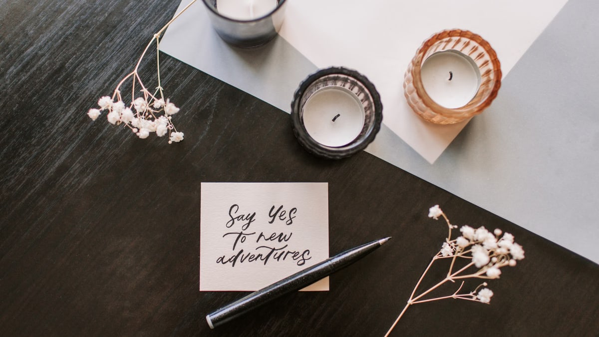 Flat lay of a motivational note, candles, flowers, and a pen on dark wooden table.